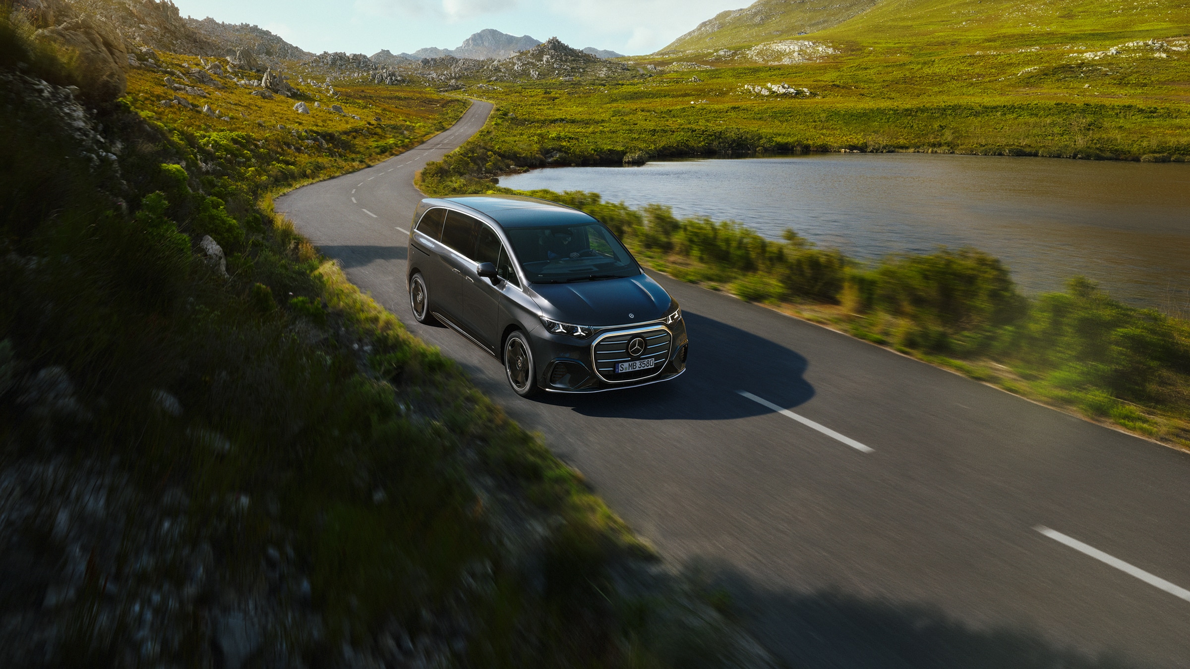 VLE | Modele de monovolume și autorulote premium | Mercedes-Benz Vans Mercedes‑Benz VLE rulând pe un drum pitoresc pe marginea unui lac, ilustrând confortul și spațiul de nivel premium.