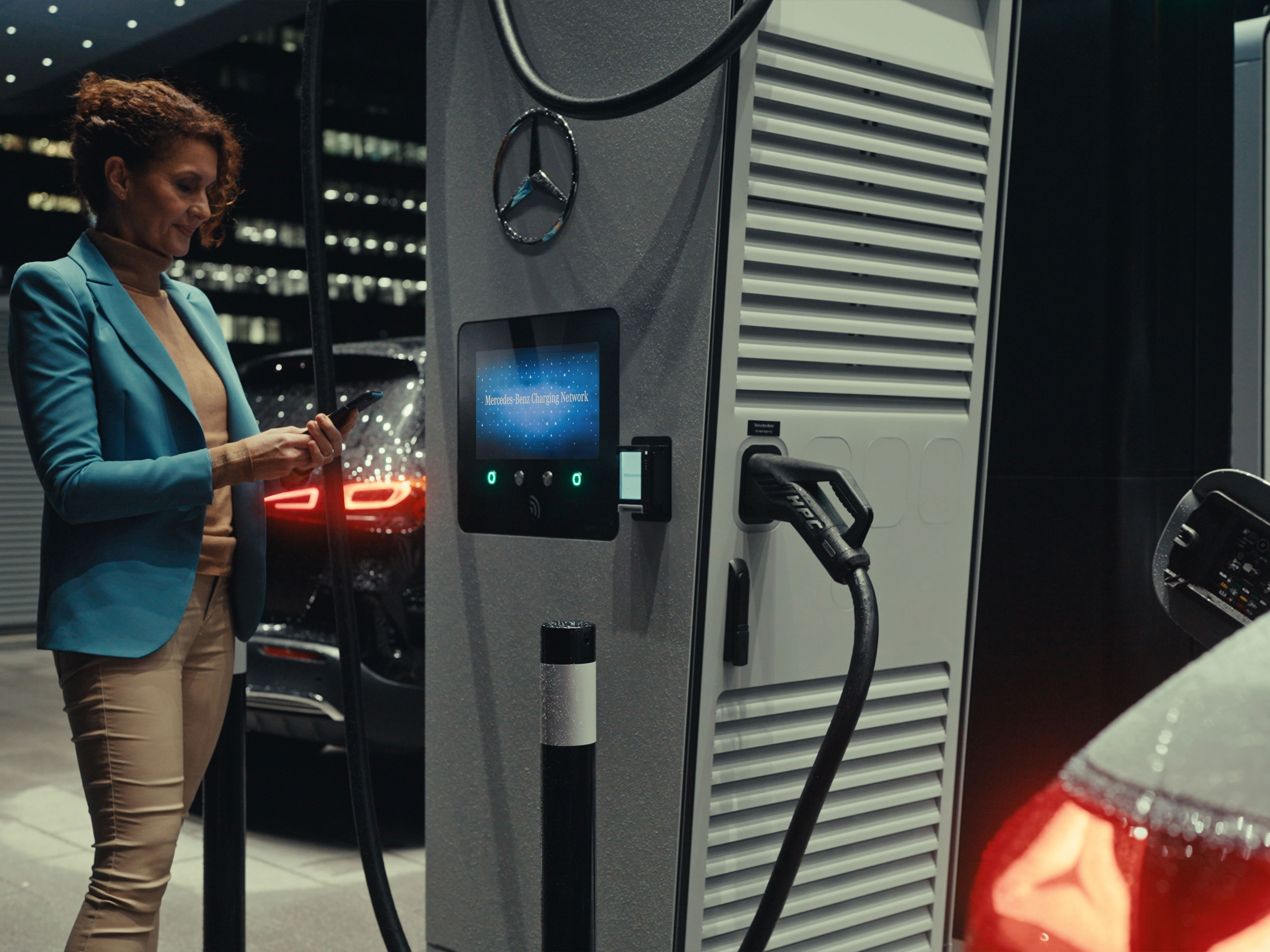 Aplicația Mercedes-Benz | Încărcare pe drum | Mercedes-Benz O femeie care ține un smartphone în mână, stând lângă o stație de încărcare.