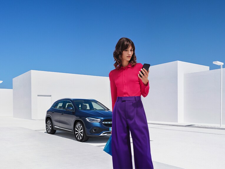 O femeie se conectează cu autovehiculul său Mercedes-Benz, folosind smartphone-ul și aplicația Mercedes me, iar cu ajutorul extraopțiunilor digitale pentru tema Remote verifică dacă sunt încuiate toate ușile autovehiculului.