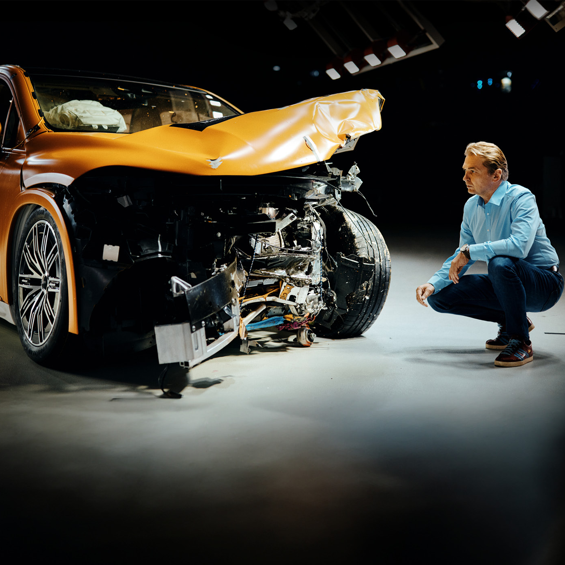 Experți în teste de impact | Siguranță | Mercedes-Benz Un expert în testele de impact examinează un autovehicul Mercedes-Benz lovit frontal, în timp ce stă ghemuit lângă ea.