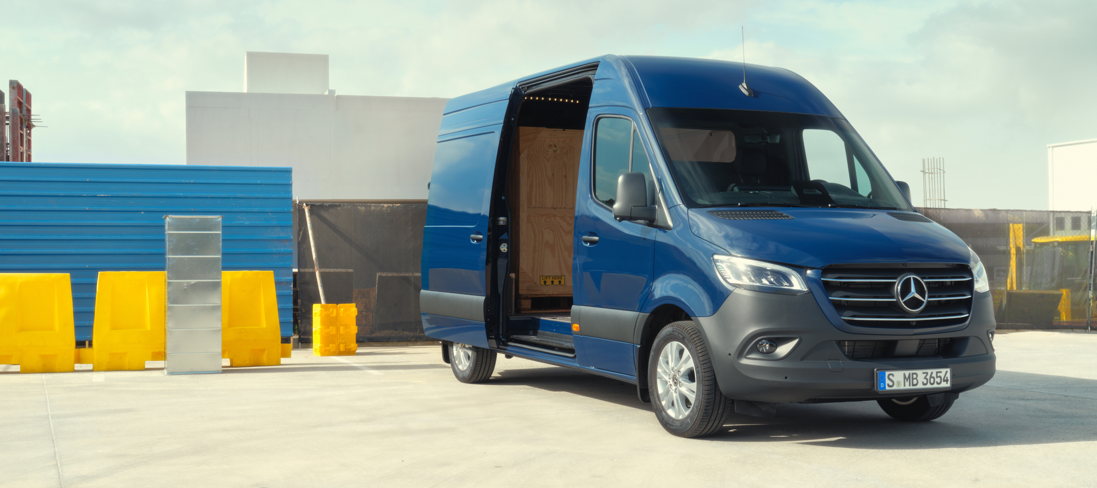 Comparație autovehicule | Sprinter furgonetă cu panou | Mercedes-Benz Modelul Mercedes-Benz Sprinter furgonetă cu panou în fața unor materiale de construcții.