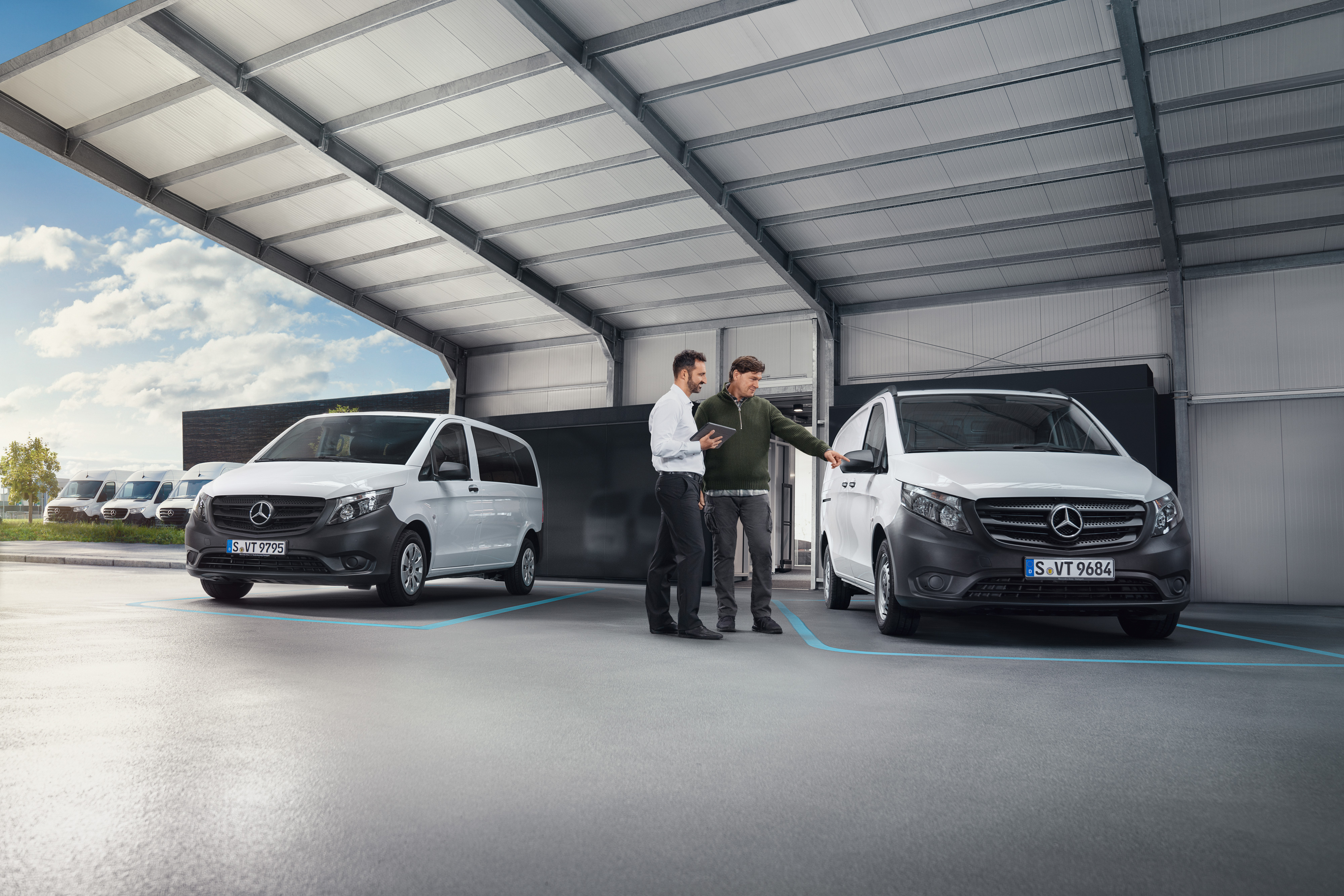 Mercedes-Benz | Comoditate | Recepție digital a clientului Doi oameni stau de vorbă într-un atelier, stând în fața a două autovehicule comerciale Mercedes-Benz albe.