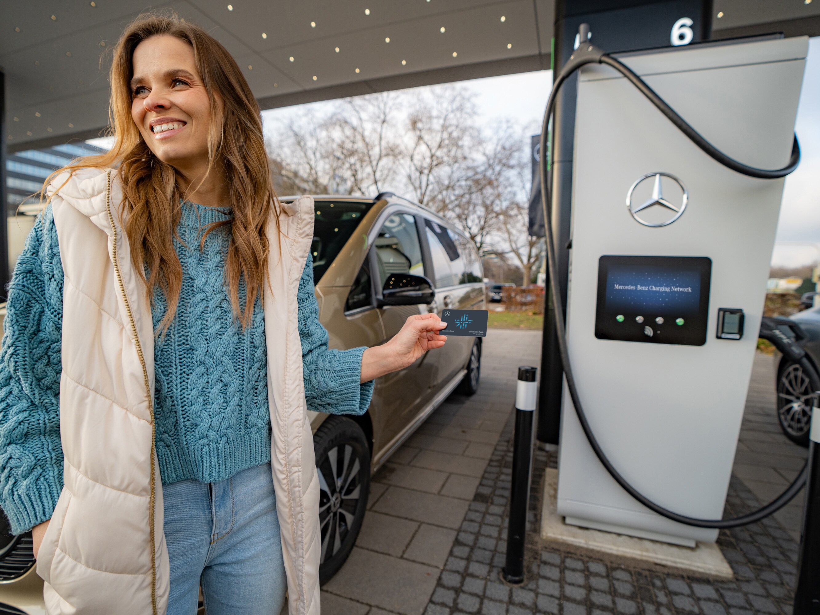 O femeie este langa o pompa de incarcare electrica. Poarta o vesta lunga gri si tine in mana un card pentru pompa