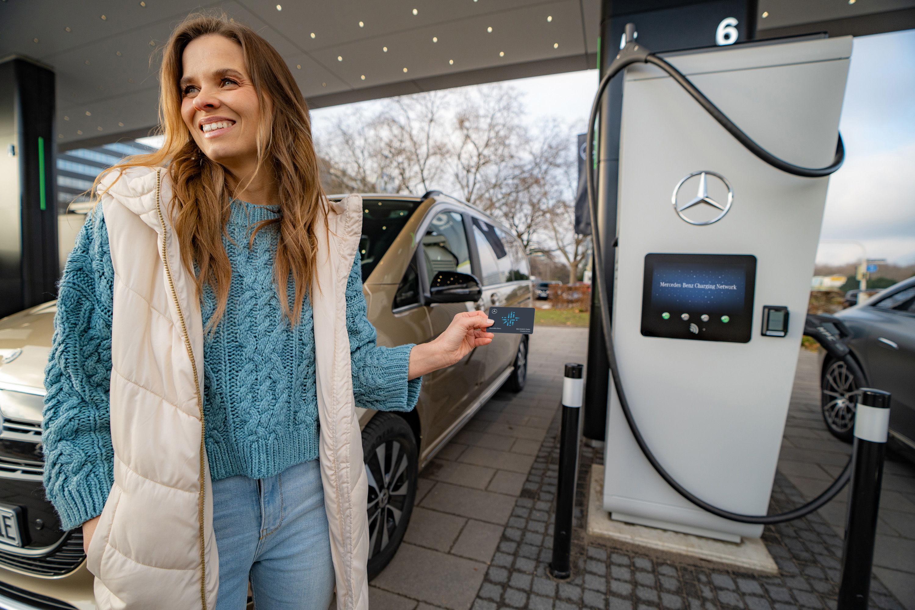 Servicii financiare | Mercedes-Benz Financial Services O femeie este langa o pompa de incarcare electrica. Poarta o vesta lunga gri si tine in mana un card pentru pompa